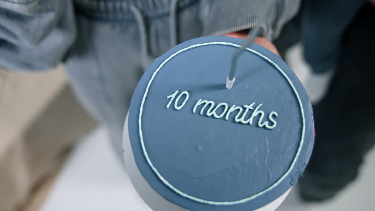 Blue cake with a sign of ten months. Unrecognized female hand lighting a candle on the cake. Man with baby stands beside. Top view.