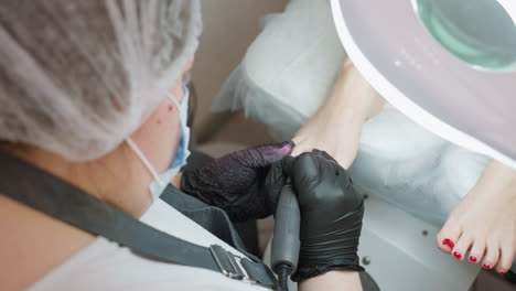 close up view of nail technician filing client toe nail using electric file under bright salon lamp, with technician in uniform and client foot with red polish resting on cushioned surface