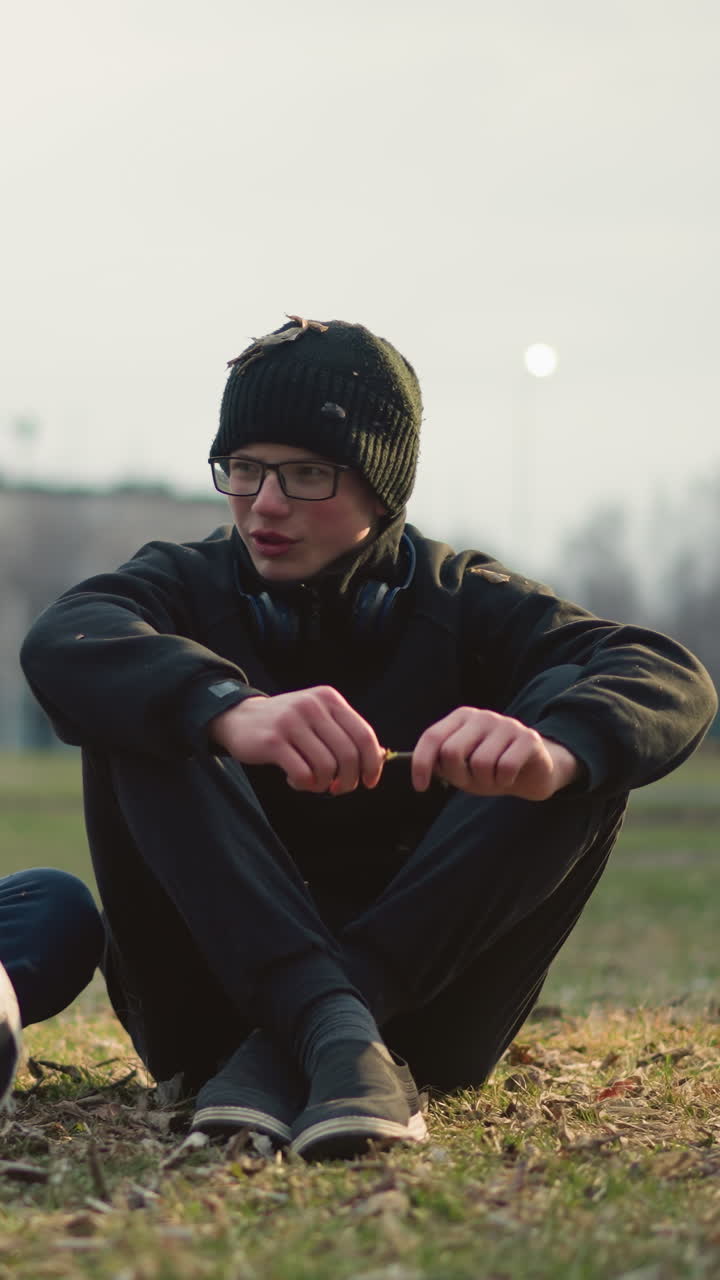 Two boys are seated on a grassy field, engaged in a discussion, the boy wearing glasses is holding a stick, attempting to cut it, while the other boy in a gray top has a soccer ball in front of him