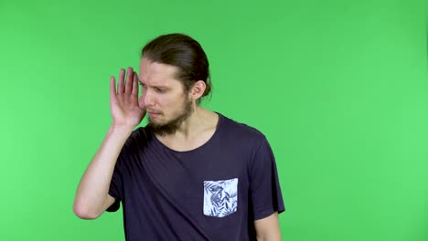 portrait of a young man holding his hand near his ear trying to listen to interesting news, expressing the concept of communication and gossip. bearded guy in the studio on a green screen. close up