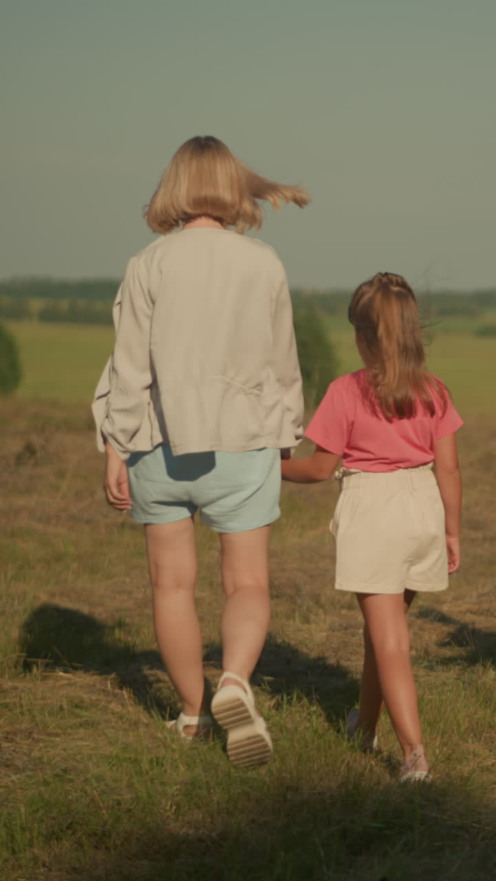 Back view of woman walking with dog on leash and mother holding her daughter s hand, strolling through farmland with lush greenery and distant trees under clear blue sky