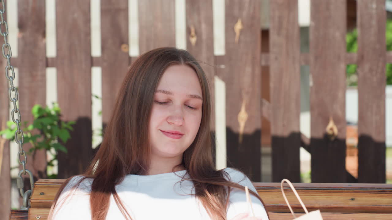 Close up of young girl sitting calmly on wooden swing in sunny backyard, she rests with arms folded, wearing white long-sleeve top, with shopping bag beside her, wooden fence in background