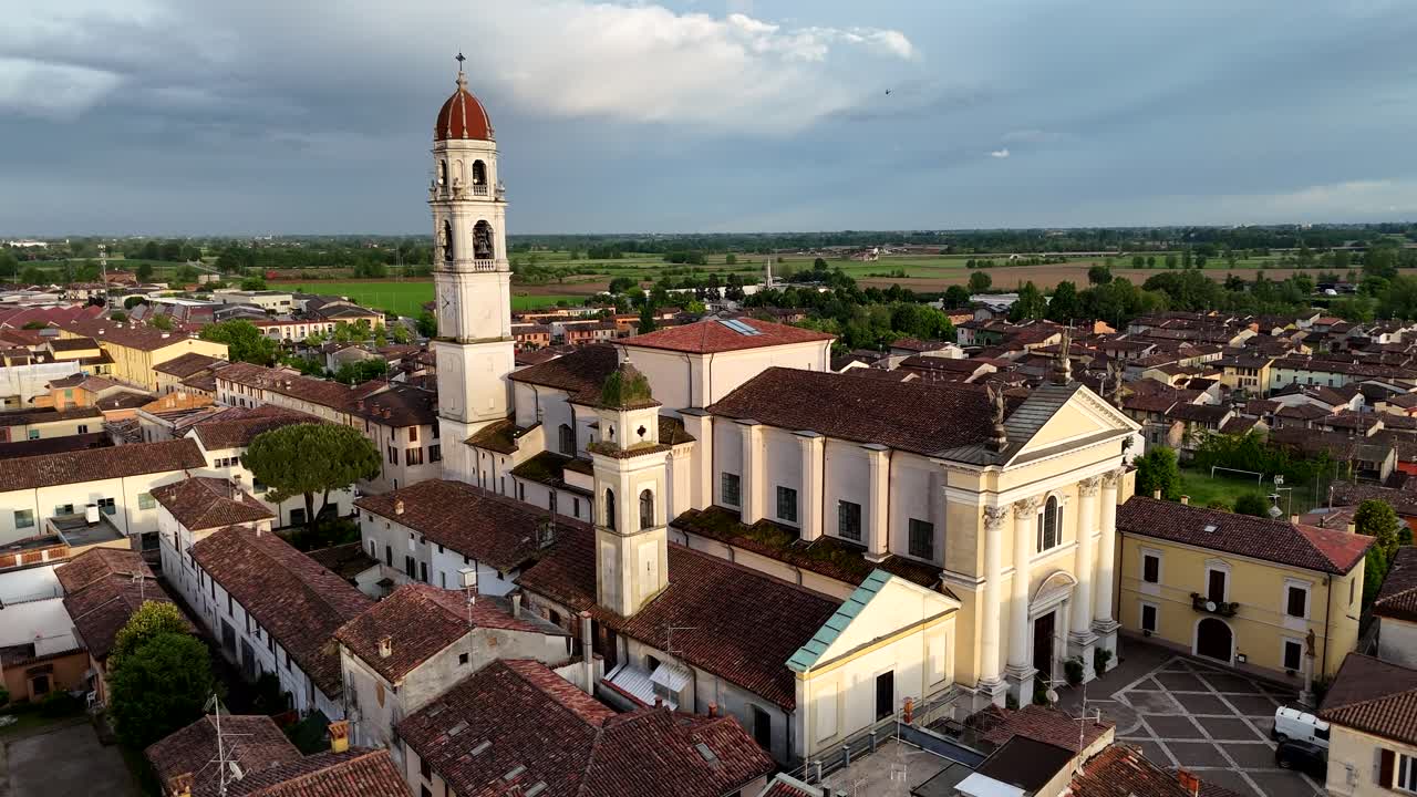 Ascending drone shot tilting down on Chiesa di Santa Maria Assunta in Pontevico, Brescia, Lombardy, surrounded by historic tiled rooftops and the rural Italian countryside stretching into the horizon
