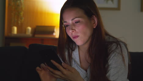 woman sitting on sofa at home at night streaming or looking at online content on mobile phone 2