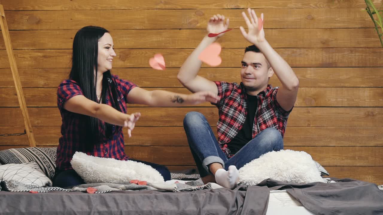 Happy enamored couple are sitting on the bed and throwing up red paper hearts and kissing while waiting for Valentine's Day. Slow motion.