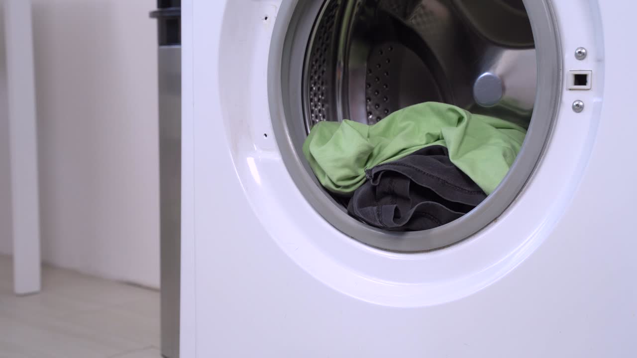 Funny little dachshund wearing blue shirt and red cap taking out uploaded laundries from the opened washing machine drum door and running away. Humor concept of dog housekeeping.