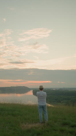 a man stands contemplatively in a grassy field by a tranquil lake at sunset. he is dressed in a white shirt, hat, and jeans, capturing a peaceful, reflective moment as the sun sets on the horizon