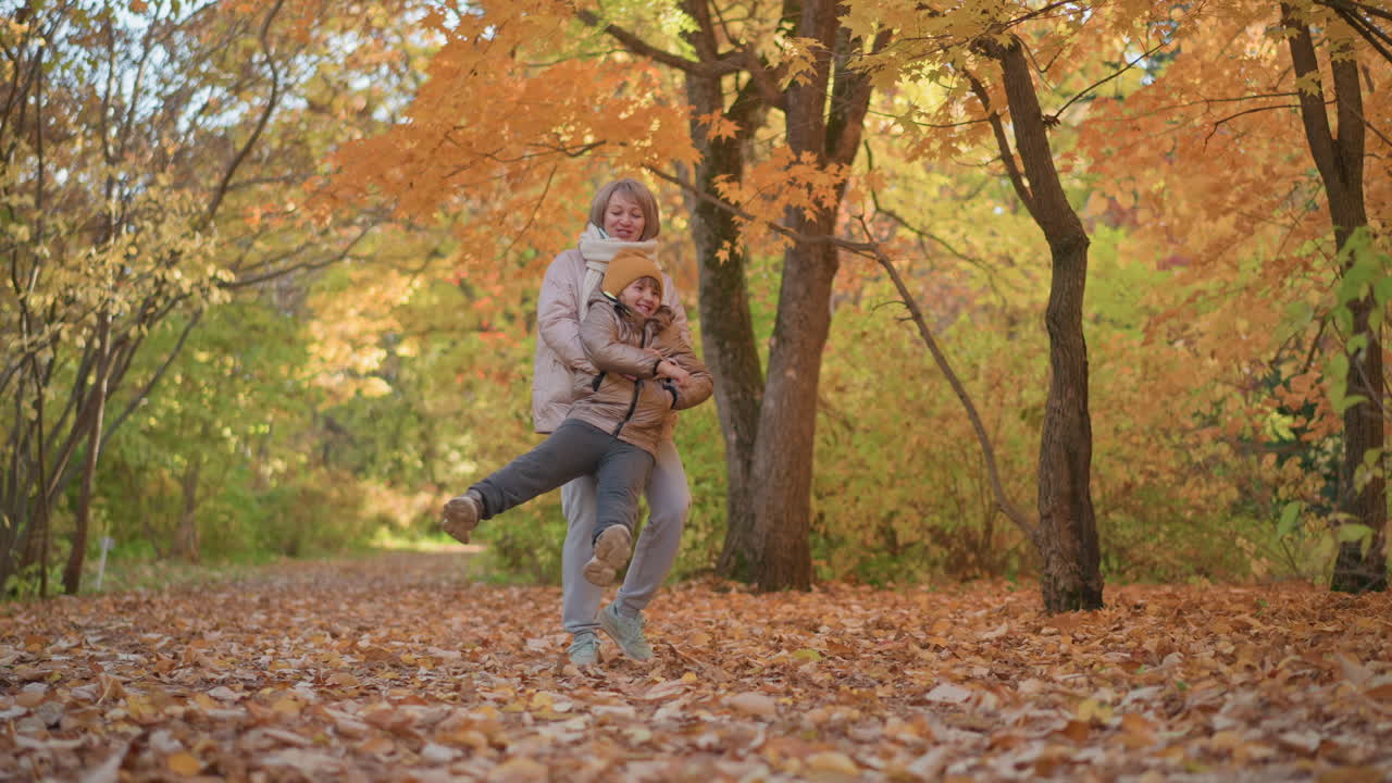 mother spins daughter around on leafy forest path during golden autumn afternoon, playful moment captured as daughter laughs and stretches legs outward beneath canopy of orange