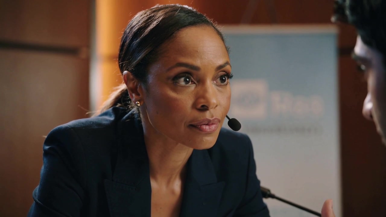 Leaning moderator wearing dark blazer and headset mic, listening at hearing table to man speaking