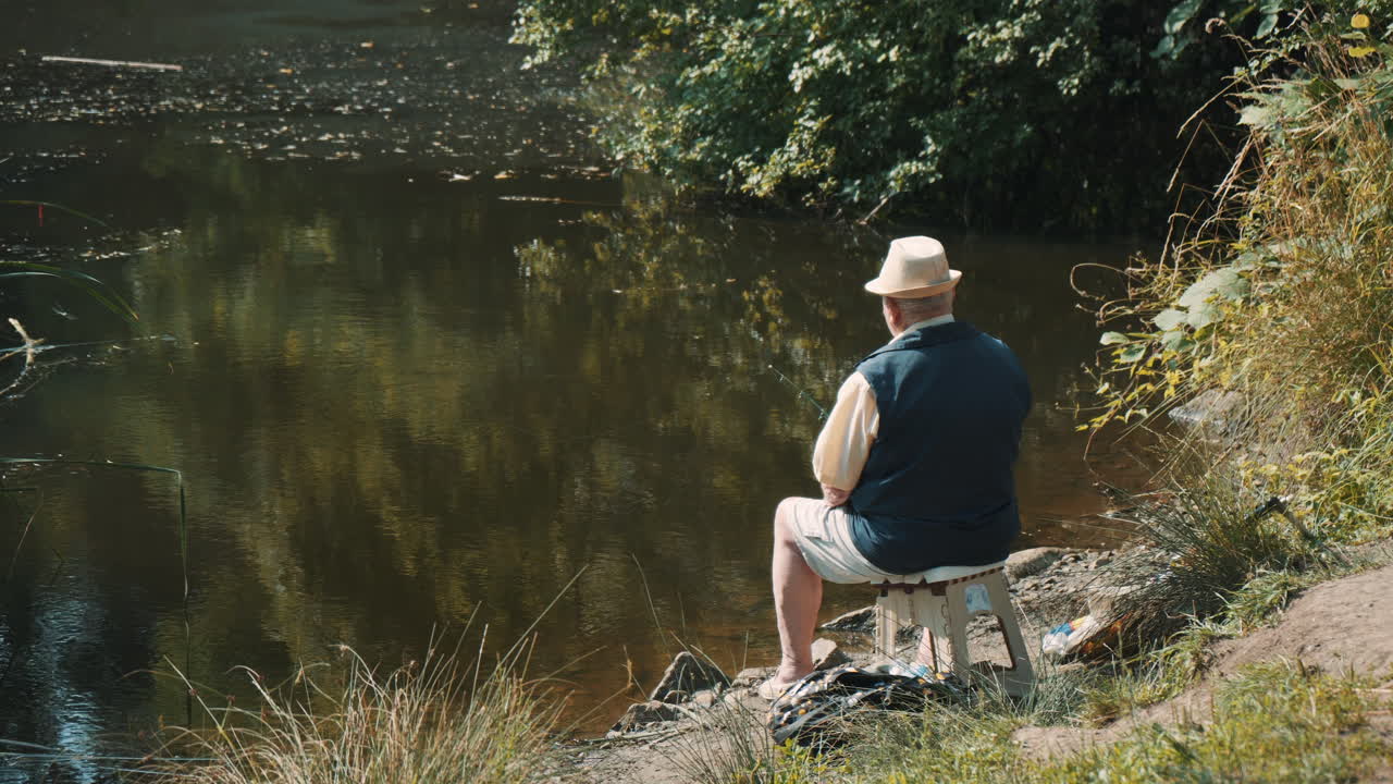 Back view of old fisherman sitting  аlone on lake shore and waiting for fish to nibble