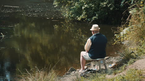 back view of old fisherman sitting  аlone on lake shore and waiting for fish to nibble