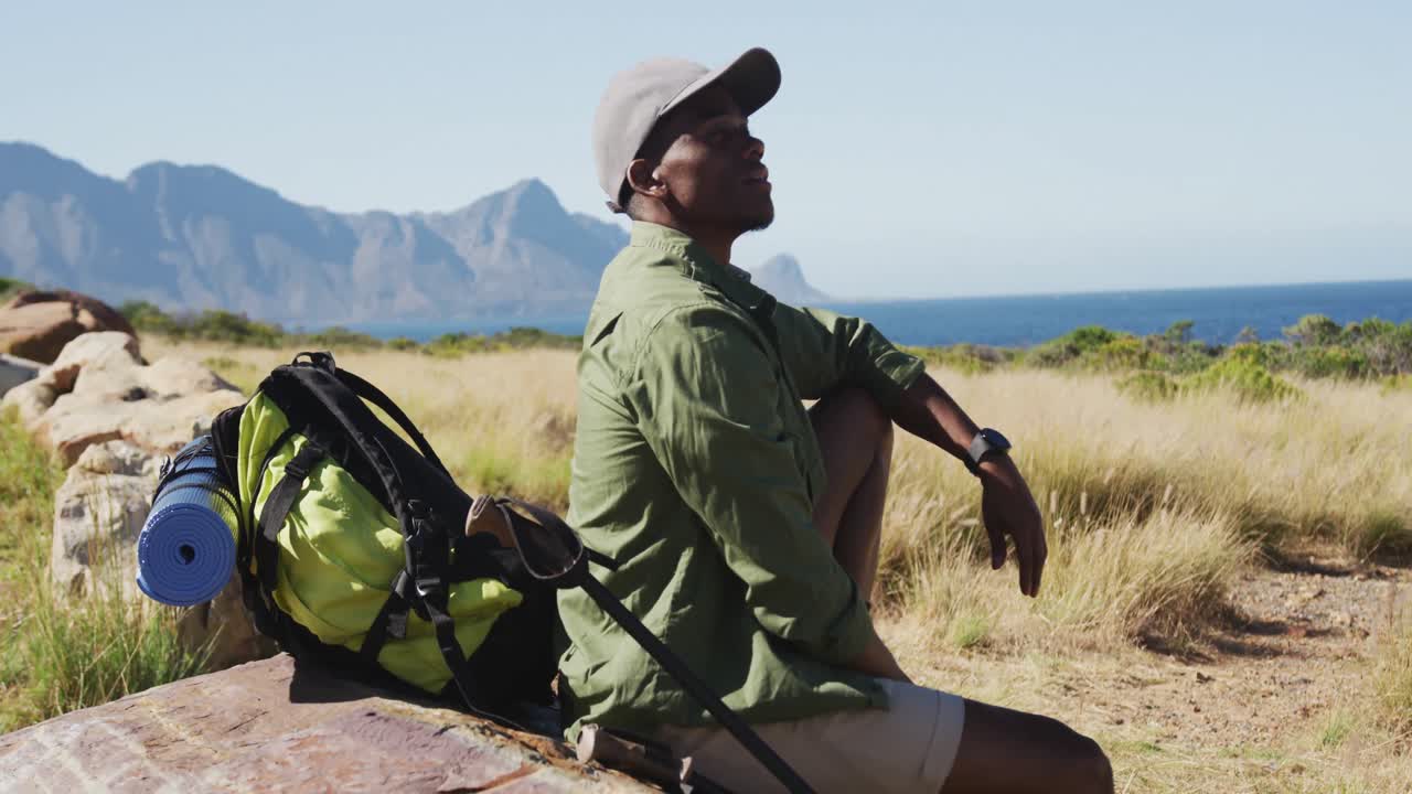 hombre afroamericano caminando en el campo por la costa tomando un descanso sentado en una roca