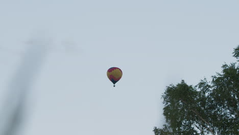Colorful hot air balloon floating high in clear sky with lush tree canopy visible in lower frame, vibrant yellow and red envelope panels contrasted against pale pastel blue background