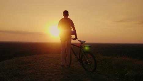 a man with a backpack and a bicycle. it stands and looks at the horizon where the sun sets. cycling and adventure