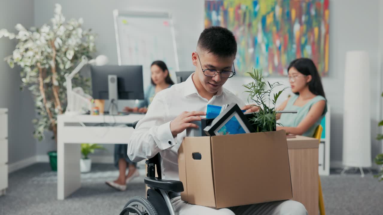 smiling, cheerful man in glasses is disabled rides wheelchair for this reason gets a promotion to another position, holds on lap a cardboard box with things, accessories, checks completeness