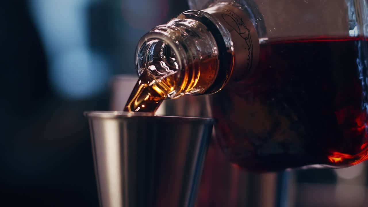 Primo piano di un distillato scuro versato in un misurino di metallo in un cocktail bar con luci soffuse.