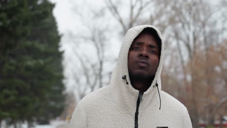 nigerian man in cozy white hoodie with fur detail chewing bubble gum, intense gaze, background of bare winter trees and park setting, providing a serene yet contemplative winter atmosphere