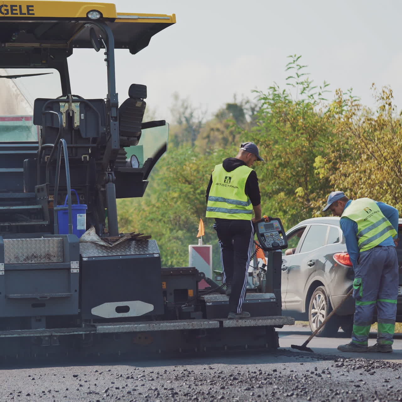 Asphalt paver machine and workers during the road construction. Male workers straighten black bitumen with special instruments on the background of traffic.