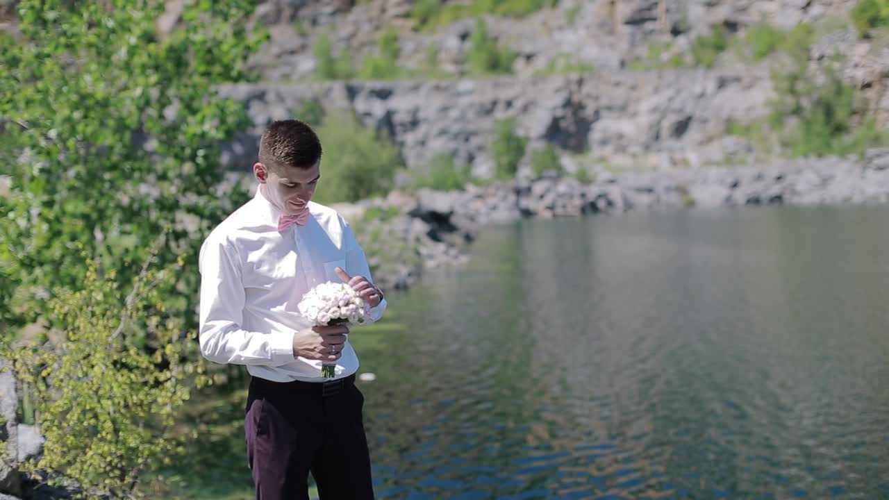 Stylish Groom Walking Alone On The Rocky Beach. Elegant stylish smiling young groom with bouquet of flowers walking alone on the rocky beach