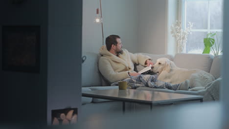 man reading on cozy sofa with golden retriever during cold winter day