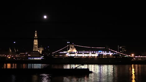 a cargo boat passing slowly under a full moon at the cityscape of antwerp