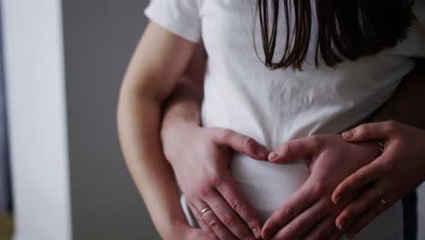 loving young husband and wife make heart sign hugging pregnant belly at home