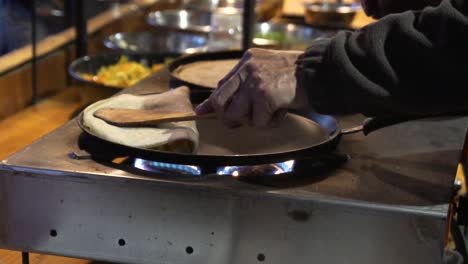 placing a pan with a pancake with bacon and vegetables on a burner in a street market