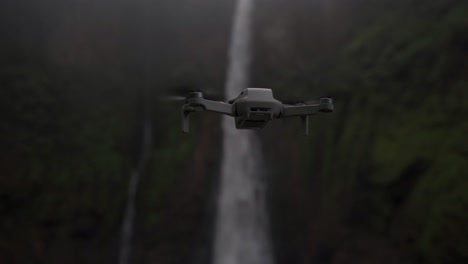 a modern drone is captured in mid-flight, hovering with a powerful waterfall cascading down a misty, green cliff face in the background