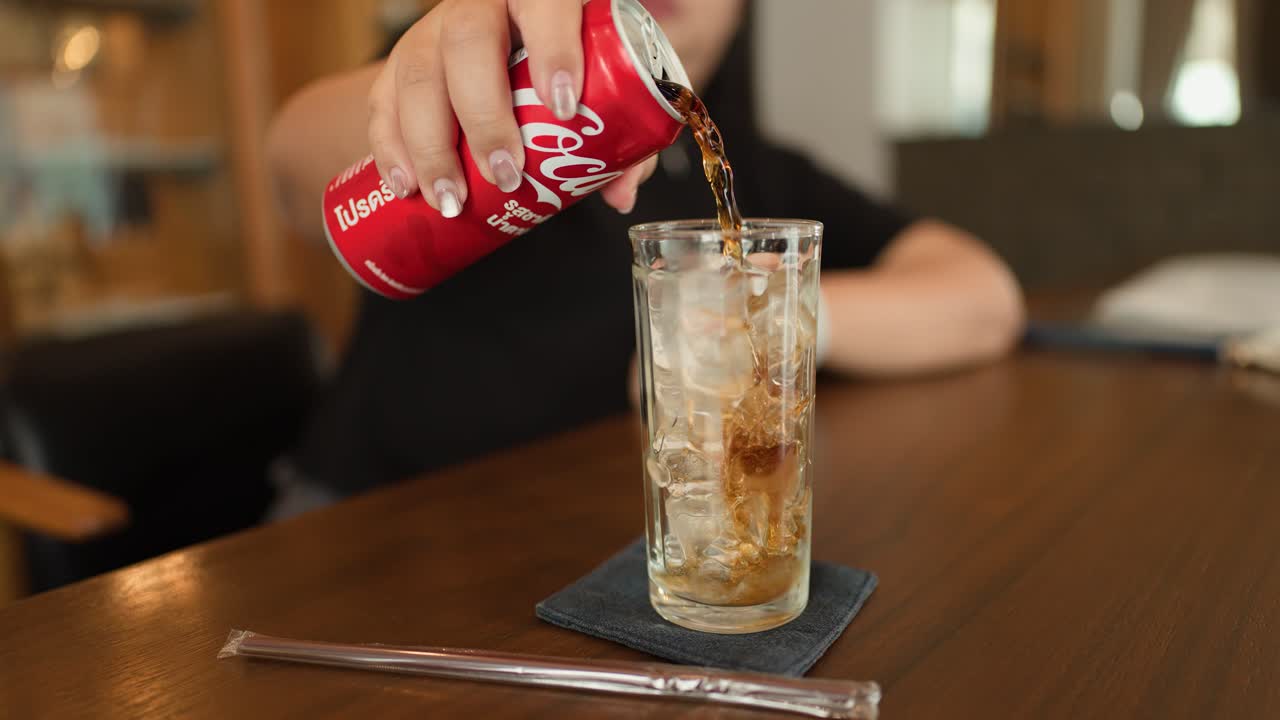 Hand pours cola into ice-filled glass on wooden table, natural lighting, relaxed dining atmosphere