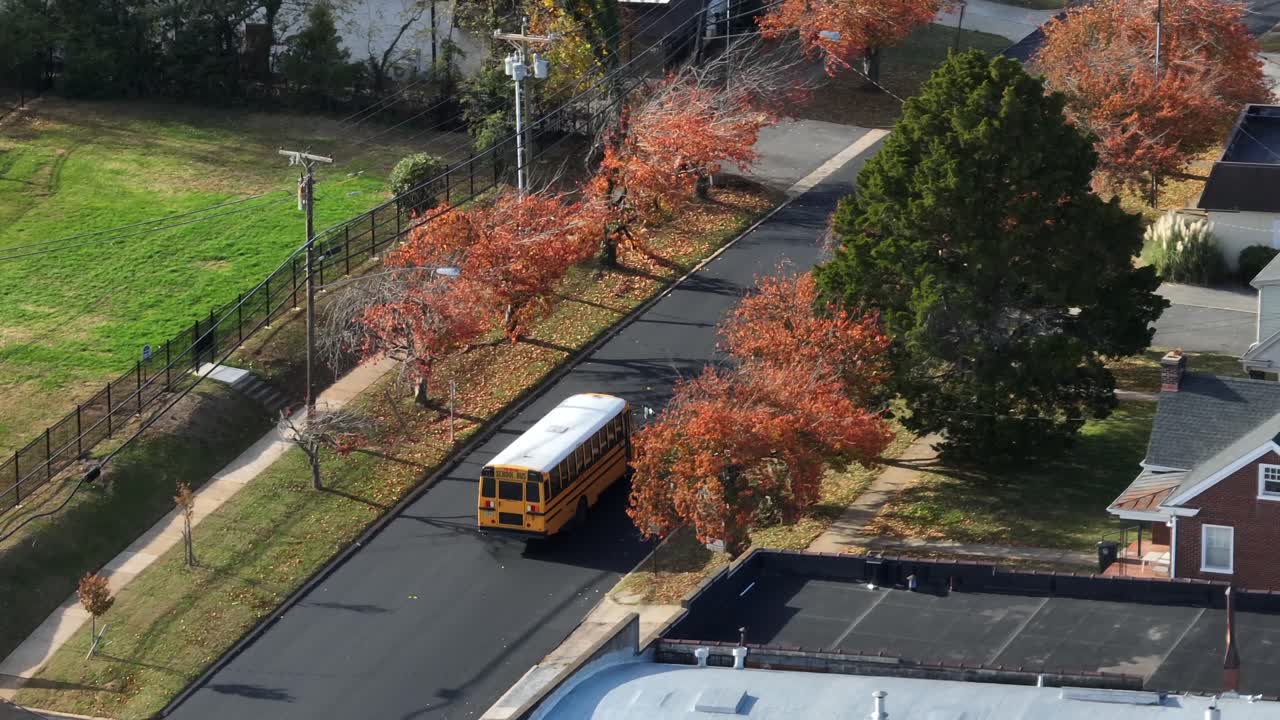 Luftfoto af gul skolebus, der kører gennem et amerikansk boligkvarter om efteråret med farverige efterårstræer, stille gader og forstadshuse under klart dagslys. Sporingsskud
