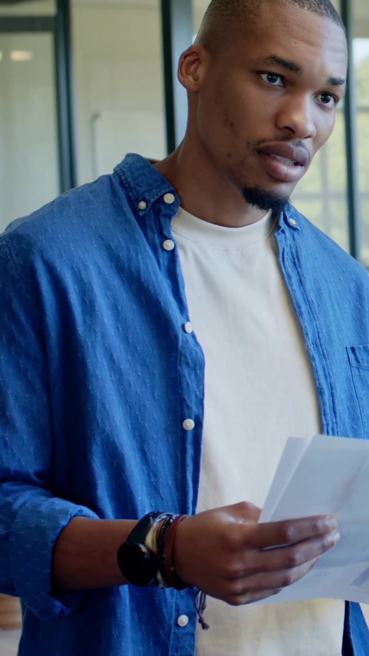 Vertical video: Scanning man in blue shirt reviewing pages after peer gesture in office, copy space