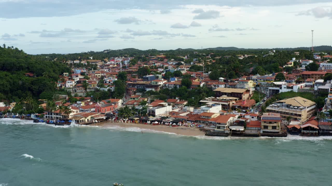 Rotating rising aerial drone shot of the famous tropical tourist beach town of Pipa, Brazil in Rio Grande do Norte during high tide with colorful buildings and exotic green foliage during the evening