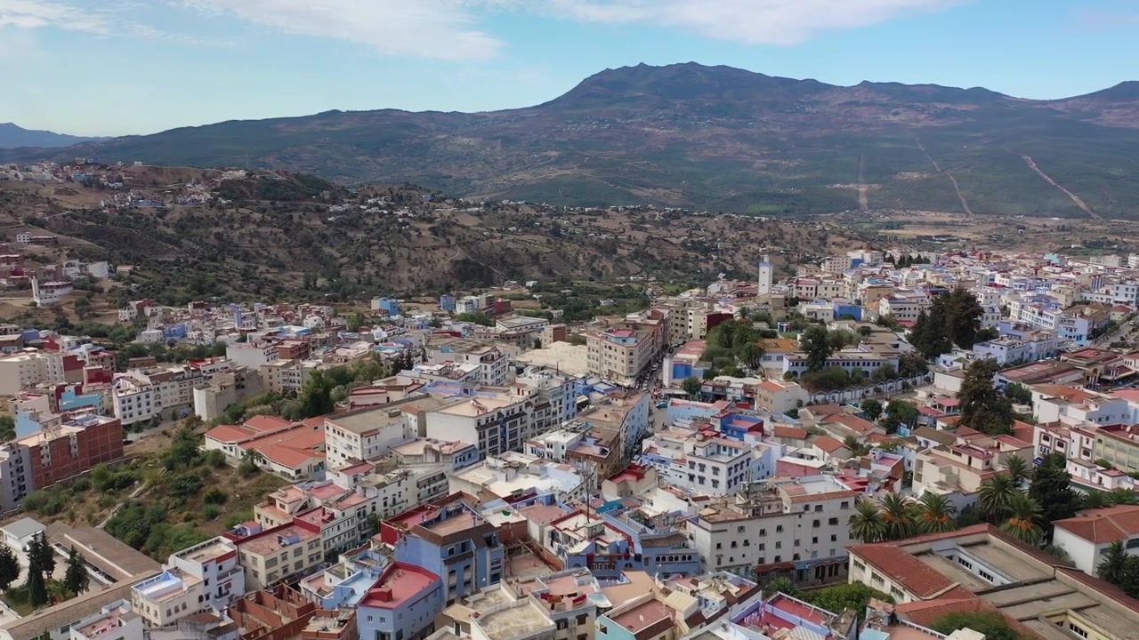 Vista aérea con dron de Chefchaouen, anidada bajo las montañas del Rif, mostrando la icónica arquitectura azul de la ciudad, el paisaje montañoso y uno de los destinos turísticos más celebrados de Marruecos.