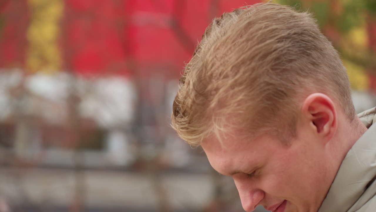 Close up of man with subtle smile walking as wind blows hair gently, autumn colors in background with soft bokeh effect showing trees and building
