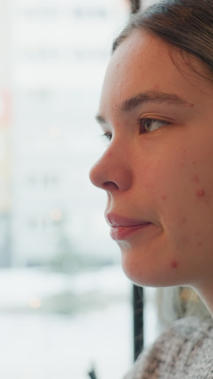 Giovane donna che sorseggia tè vicino alla finestra, sfondo urbano innevato visibile attraverso il vetro, maglione accogliente, vapore che sale dalla tazza, occhi chiusi, atmosfera da bar adattata per la casa, momento di una studente o di un freelance, inverno in città