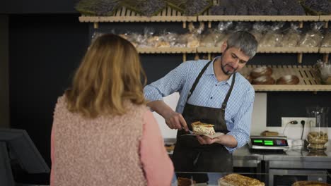 Happy-bakery-salesman-packing-a-piece-of-pie-on-paper-for-a-female-customer-while-they-have-a-casual-conversation