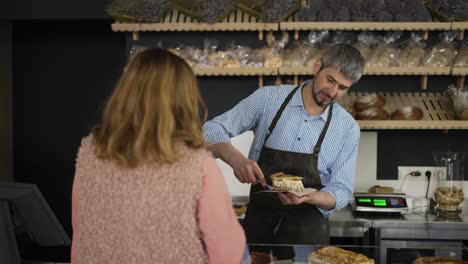 feliz vendedor de panadería empaquetando un pedazo de pastel en papel para una cliente femenina mientras tienen una conversación casual