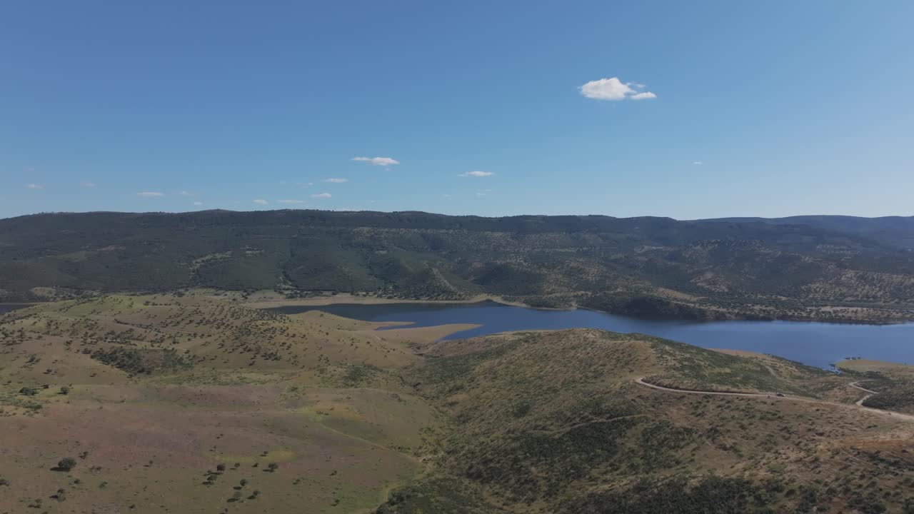 Drone flight over the Guadiana River and the Cíjara Reservoir in Extremadura. A sideways pan reveals blue waters, sun reflections, a bridge crossing the river, and a chapel under a clear sky.