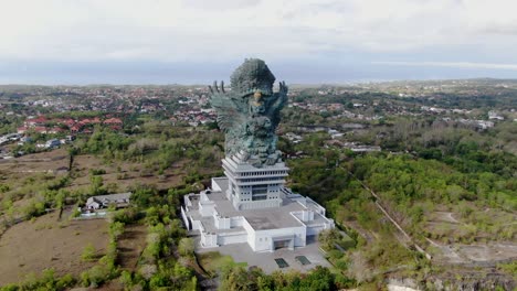 massive statue of garuda winu kencana near small town of bali island, aerial orbit view