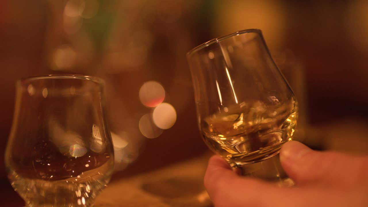 Hand swirls whisky glass in cozy, warmly lit distillery tasting room, shallow depth of field