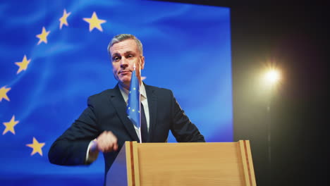 mature politician giving encouraging speech from rostrum pan around view of middle aged male in suit gesticulating and speaking convincingly with audience while standing on podium against european flag