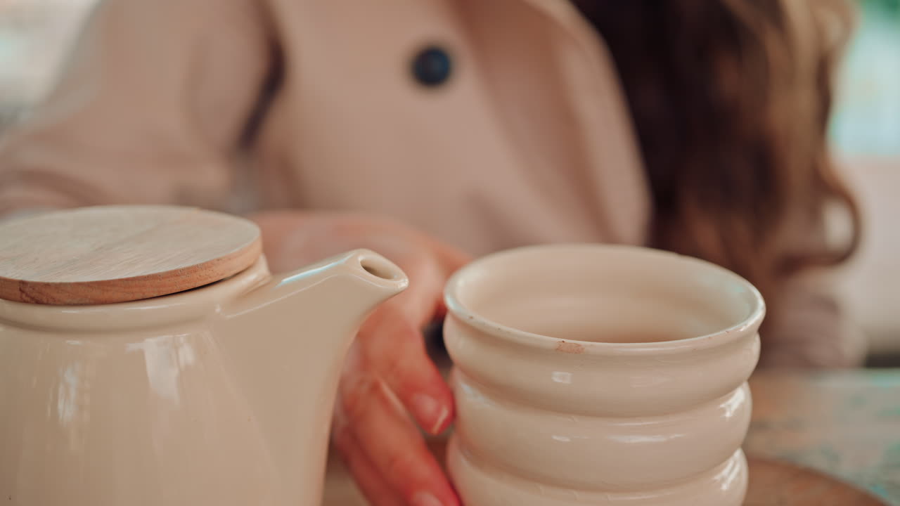 Close up of a woman pouring hot water from a ceramic teapot into a textured mug at an outdoor cafe, with soft daylight and warm tones