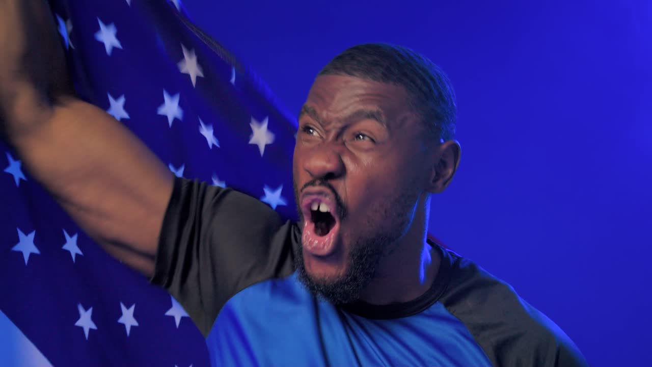 Afroamerikansk mand råber, vikler og vifter med et flag, jubler i et blåt studie iført en blå skjorte.