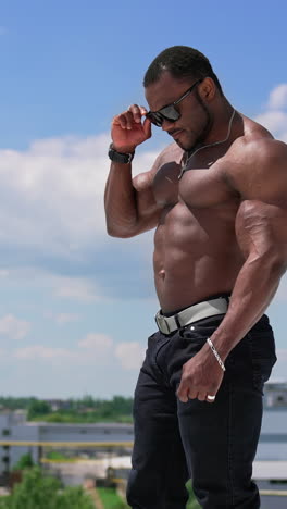 portrait of handsome african sportsman outdoors. cheerful shirtless athlete with strong trained muscles standing on the hill against blue sky. vertical video