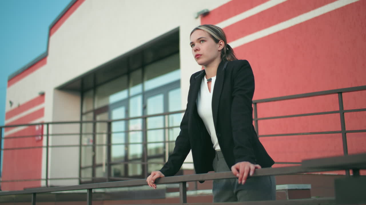 Female CEO in black blazer leans confidently on iron rail outside modern office building, displaying leadership, strength, and poise under clear sky, blending elegance with authority in urban setting
