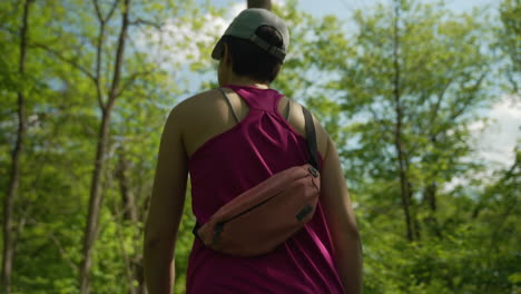 following woman hiking through the woods with a small fanny pack around her back and wearing pink tanktop