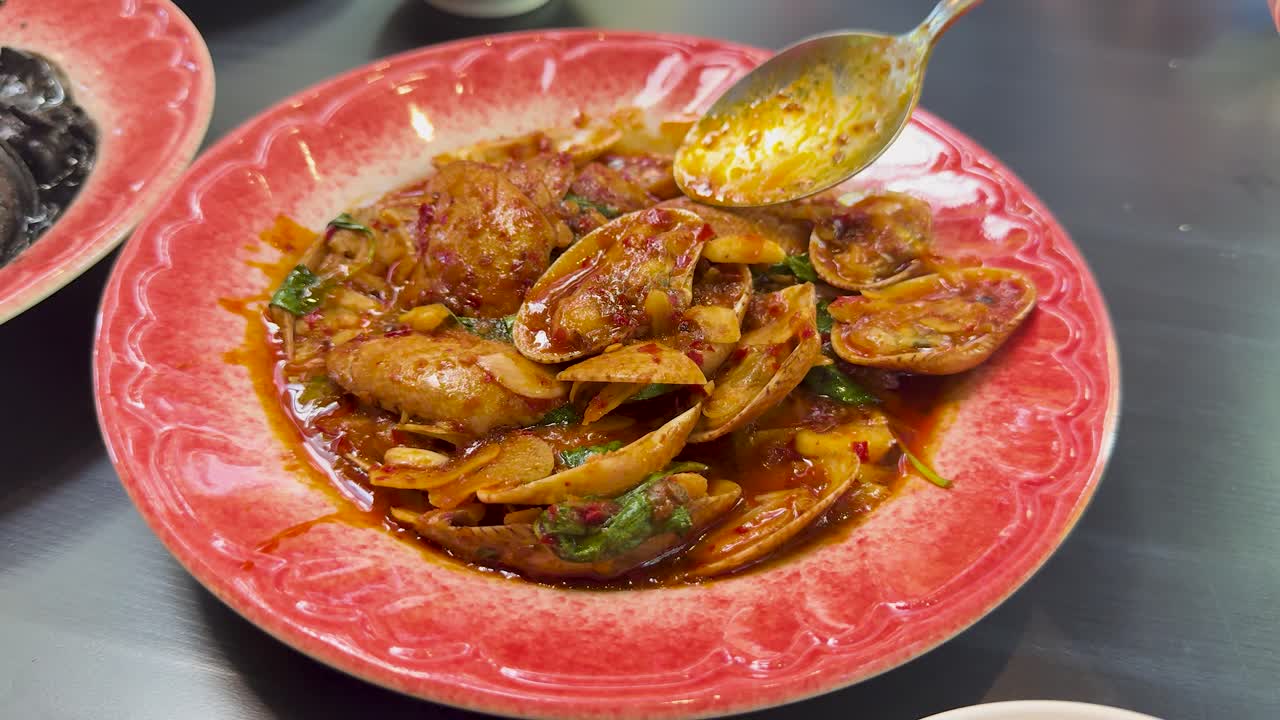 A hand uses a spoon to serve spicy clams cooked in chilli paste, presented in a vibrant red bowl under bright, even lighting on a metal surface