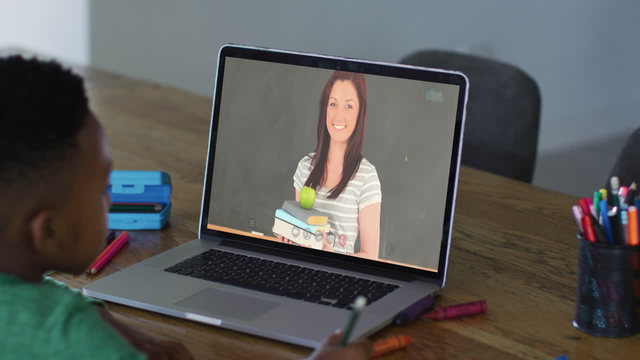 niño afroamericano haciendo la tarea mientras tiene una videollamada con una maestra en una computadora portátil en casa