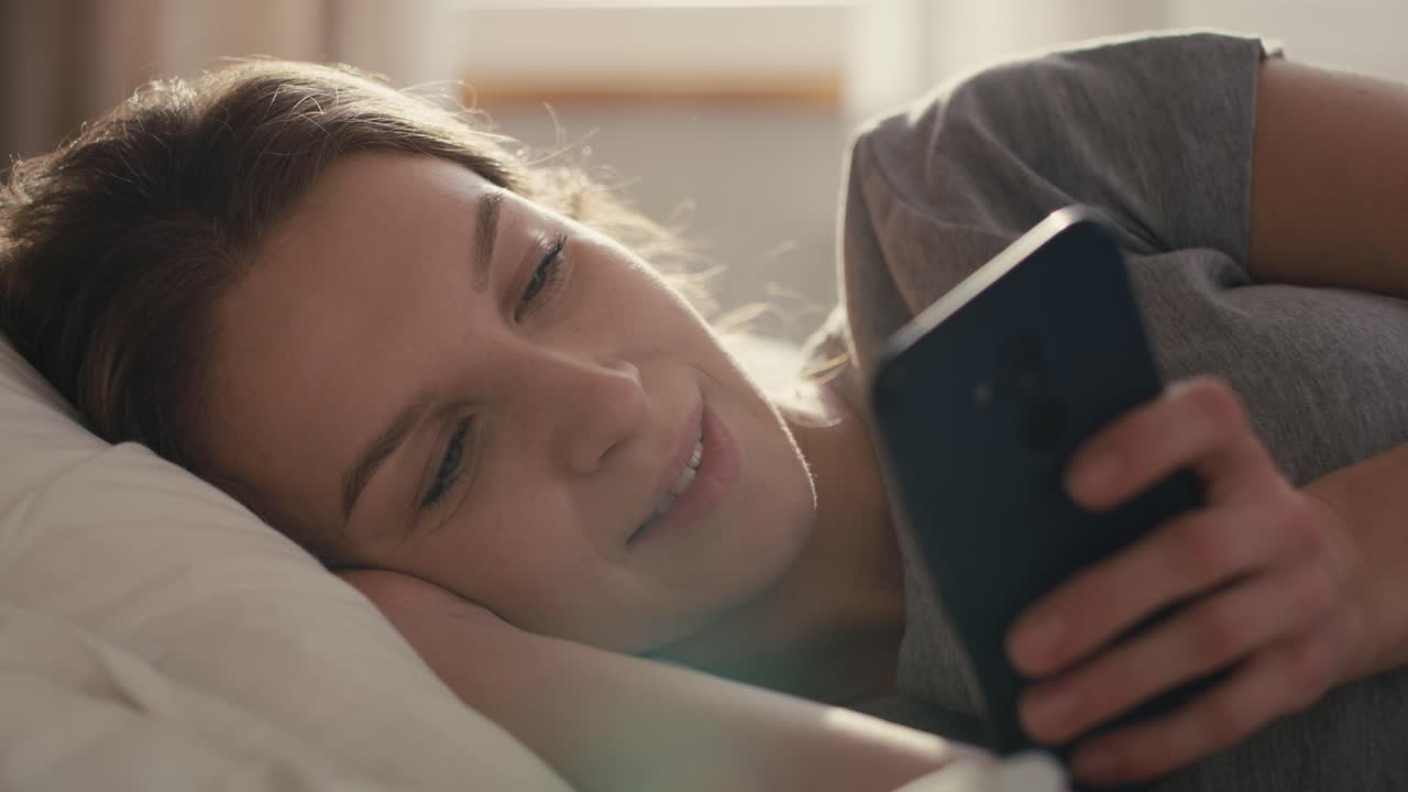 Kaukasisk kvinde ligger i sengen om morgenen og bruger mobiltelefon