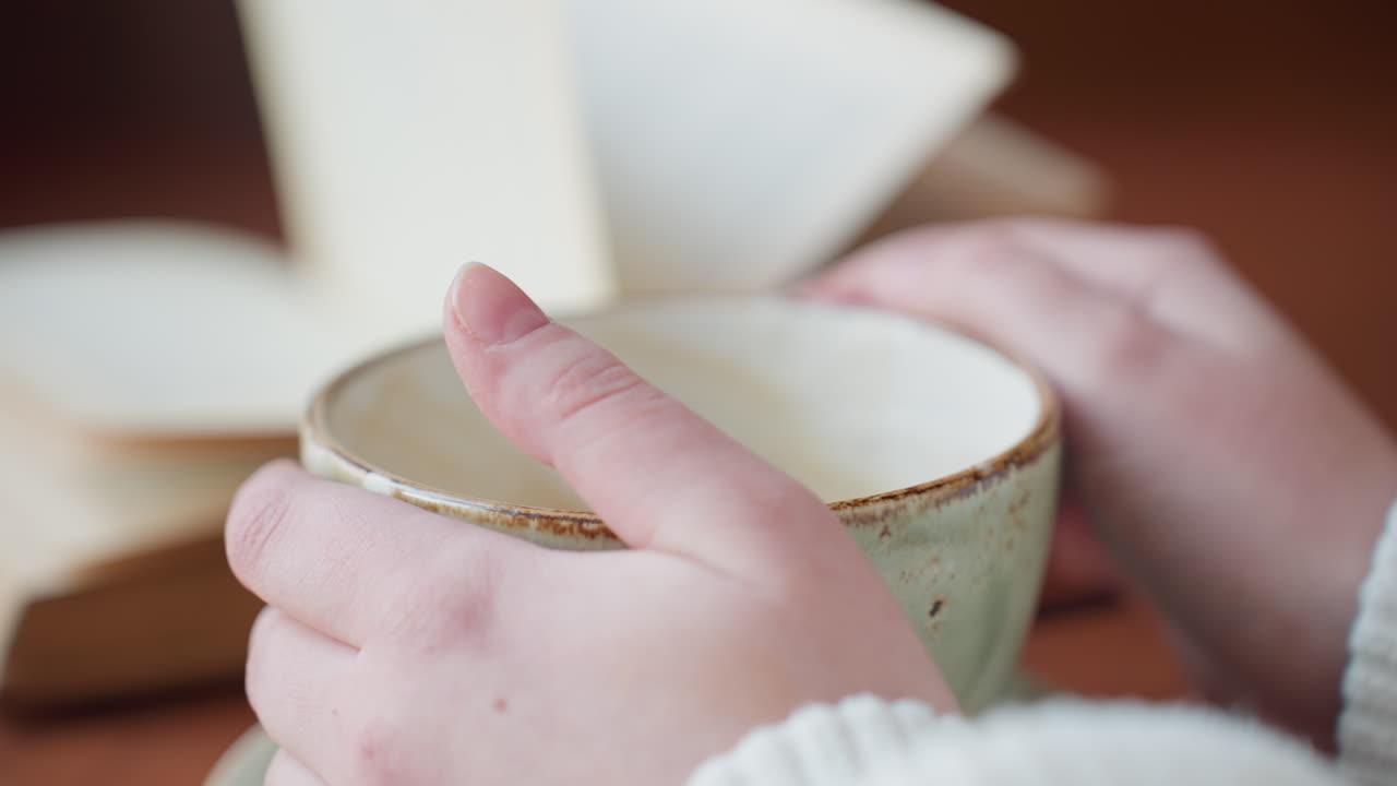 close up hand stirs coffee in ceramic cup then drops spoon and lifts cup while blurred book pages flap in breeze creating calm indoor moment with warm tones and relaxed atmosphere